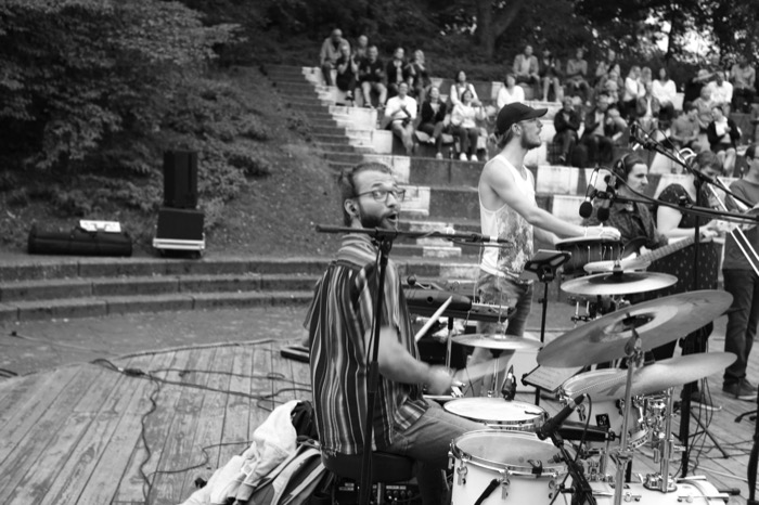 Drummer in a striped shirt singing mid-beat on an open-air stage, bandmates and tiered audience behind