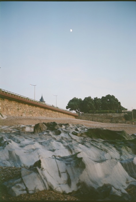 Fissured grey rock shelf below a stone sea wall and promenade, half moon in a pale evening sky