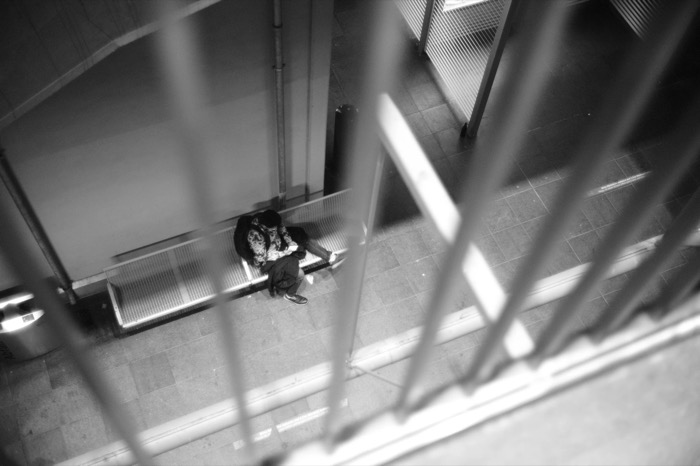Shot down through blurred railings: a person huddled alone on a bench in an empty covered walkway