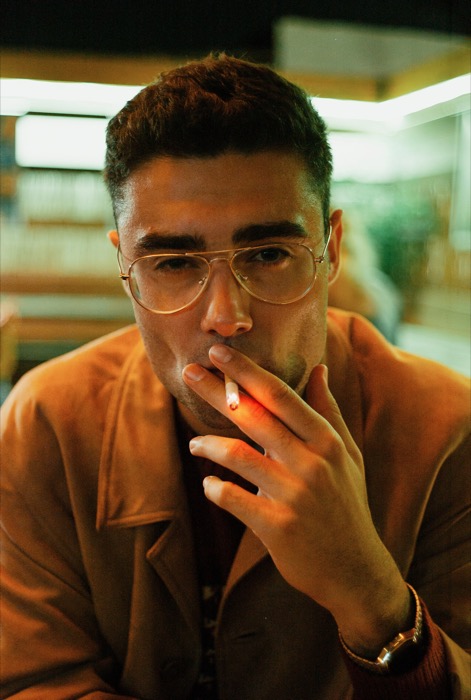Young man in glasses and a light coat smoking a cigarette indoors, staring straight at the viewer