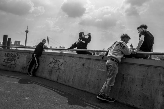 Four teenagers leaning on a graffiti-covered riverside wall, a tower and cable bridge on the skyline