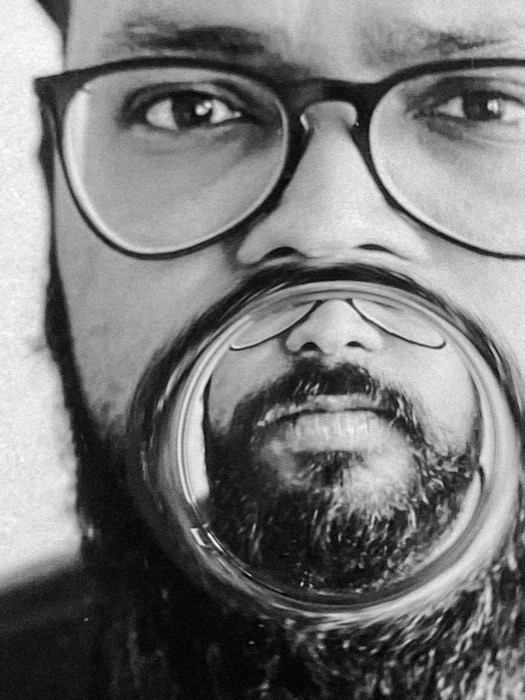 Close-up of a bearded man in glasses, his mouth and beard distorted through the base of a glass