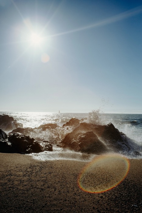 Waves bursting over dark rocks at the shoreline under a low sun, a sailboat on the horizon