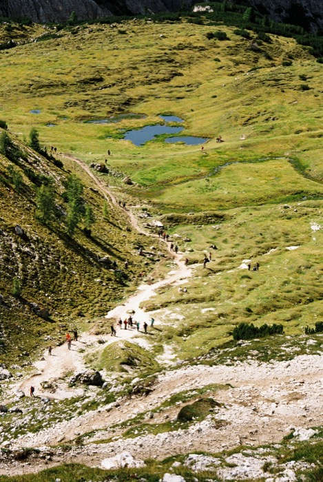 Hikers strung out along a pale gravel path winding through green alpine meadow past small pools, seen from above
