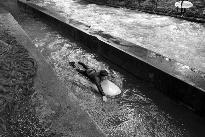 Surfer lying on a board paddling in the shallow channel beside a concrete river weir
