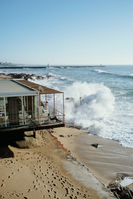 Wave exploding against rocks beside a glass beachfront pavilion where one person sits on the deck