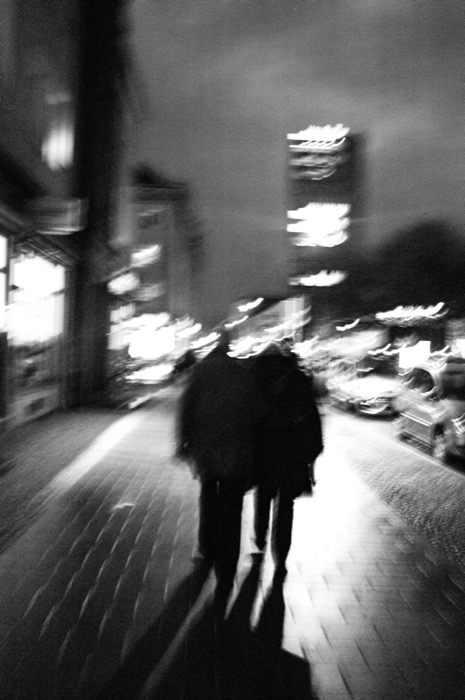 Two figures walk away down a night street, blurred streaks of shop lights swirling around them