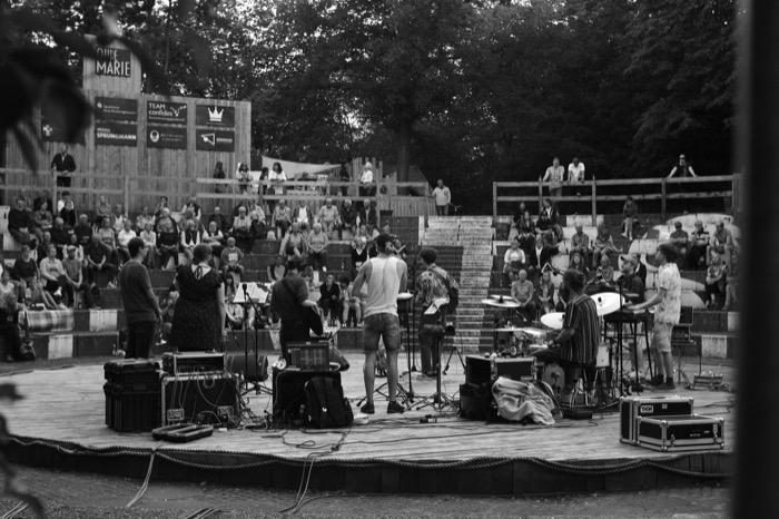 Band playing on a circular open-air stage as an audience watches from concrete tiers under trees