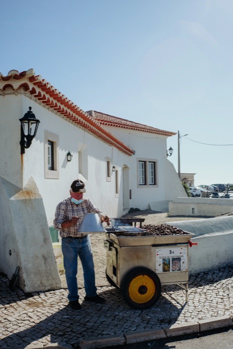 Vendor tending a chestnut roasting cart on a cobbled street beside a whitewashed tiled house
