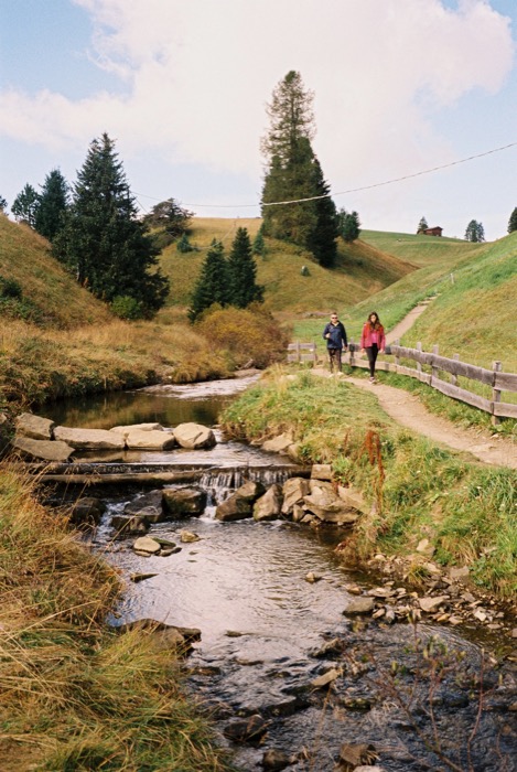 Couple walking a fenced path beside a small stream stepping over rocks in a grassy alpine valley