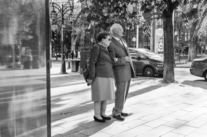 Elderly couple arm in arm in coats, waiting to cross a tree-lined city boulevard