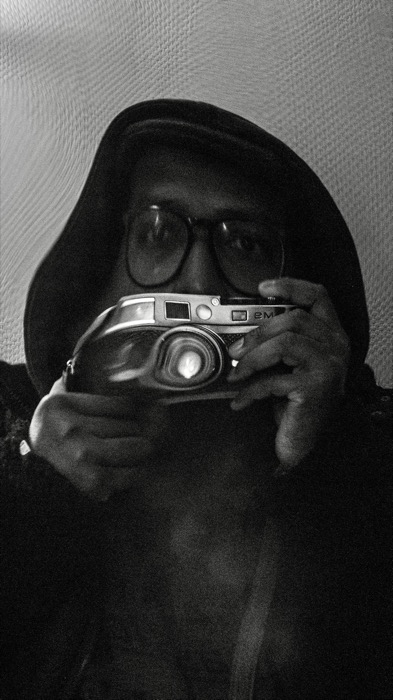 Man in a hood and glasses holding a rangefinder camera to his chest, face half-covered