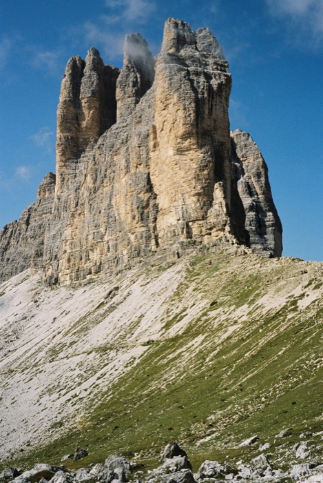 Sheer pale rock tower above a green and stony alpine slope under deep blue sky