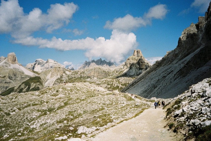 Hikers strung out along a gravel mountain path below limestone spires and towering clouds