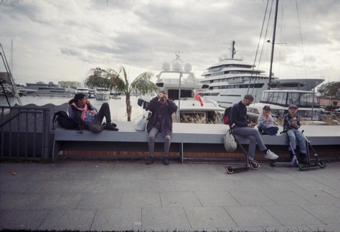 People resting on a low harbour wall in front of moored yachts under a grey sky