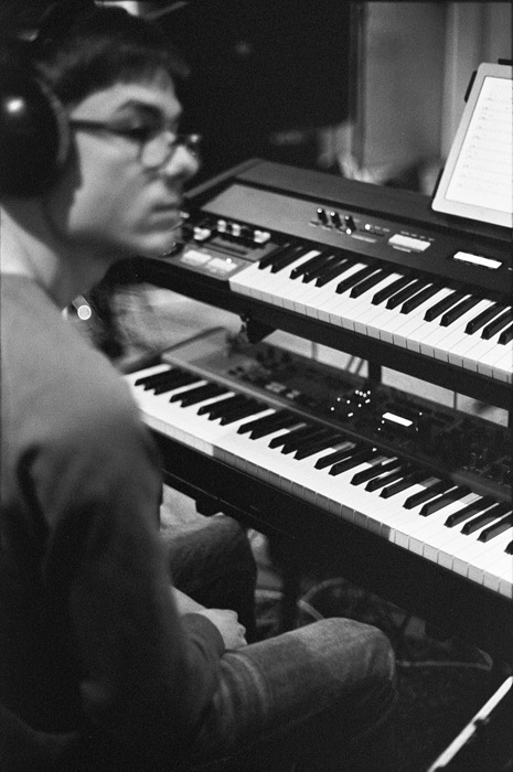 Musician in headphones and glasses seated at a stack of two synthesizers in a dim studio