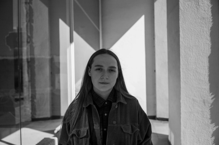 Long-haired young person in a corduroy jacket standing between glass and a sunlit concrete wall
