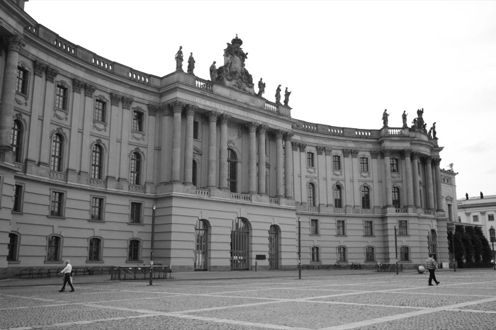 Two men crossing an empty cobbled square before the curved colonnade of Humboldt-Universitaet