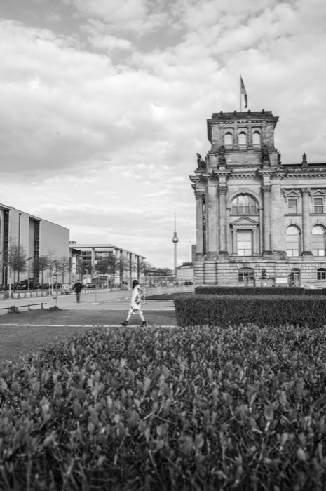 Woman in a patterned coat walking past clipped hedges below the Reichstag, TV tower in the distance