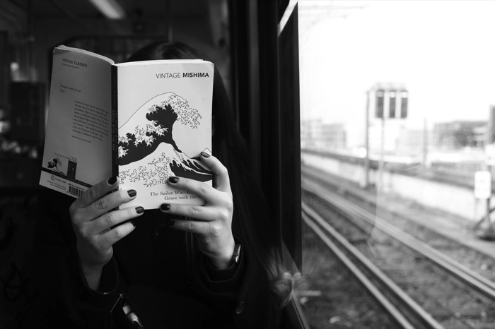 Woman on a train holding a Mishima paperback over her face, rails running past the window