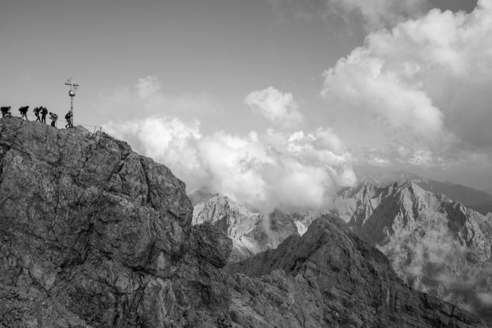 Hikers gathered at a metal summit cross on a rocky peak above clouds and jagged ridges