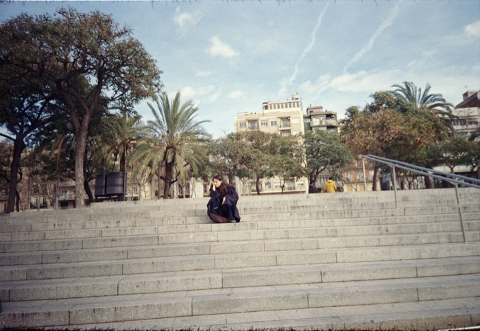 Lone figure in a dark coat sitting on wide stone steps, palm trees and apartment blocks behind