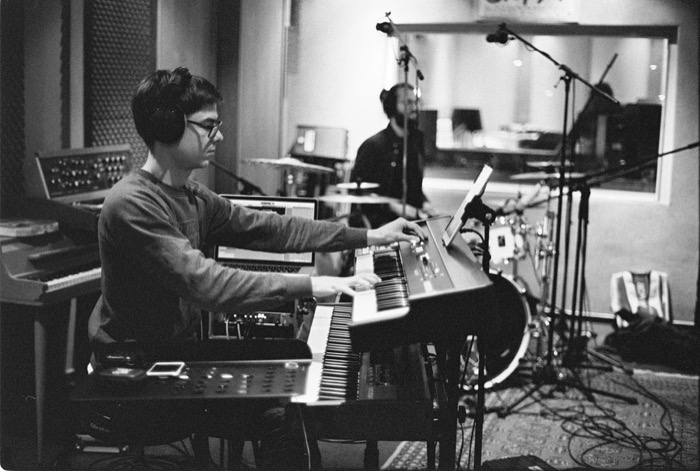 Keyboard player in headphones and glasses leaning into a synth in a recording studio, drummer behind him