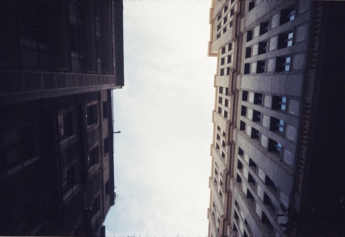 Looking straight up between two tall facades, one in shadow, one in patterned concrete, to a pale sky