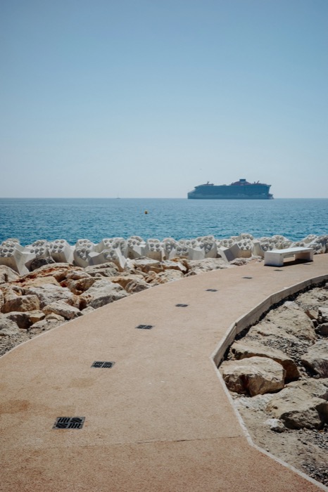 Empty curving seafront promenade with a bench, concrete breakwater blocks and a cruise ship on the horizon