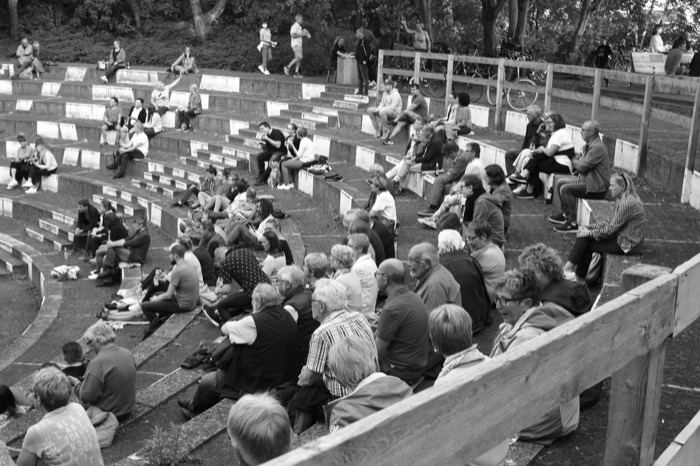 Audience sitting on the concrete tiers of a wooded open-air amphitheatre, bikes parked at the top