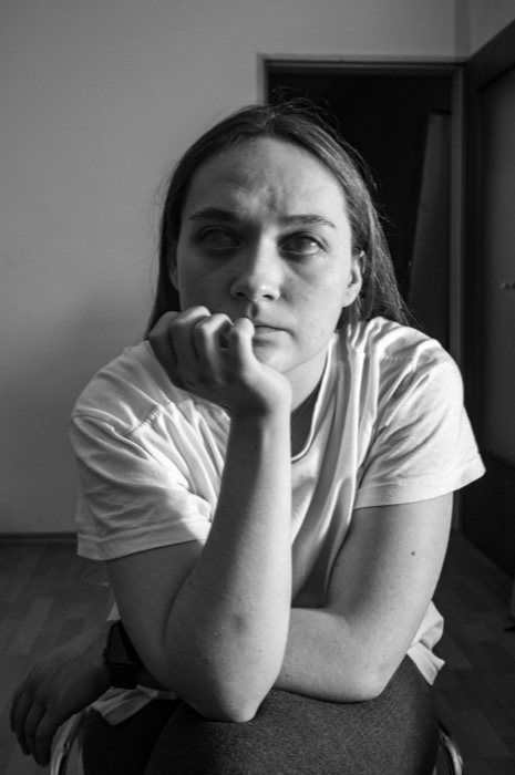 Young woman in a white t-shirt resting her chin on her fist, frowning into the camera in a bare room