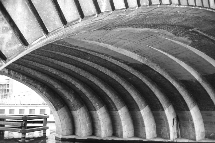 Underside of a stone bridge arch, ribbed vaults curving over the water in strong light and shade