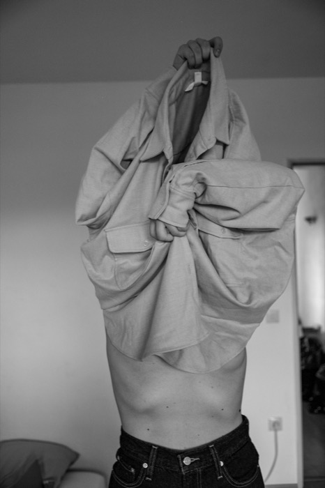 Bare-chested person pulling a shirt up over their head in a bedroom, face hidden by the fabric