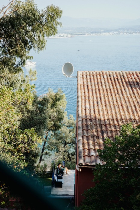 Terracotta tiled roof with a satellite dish, seen through trees above a bay, a person sitting on the terrace below