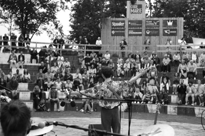 Musician seen from behind spreading both arms to a seated open-air amphitheatre crowd