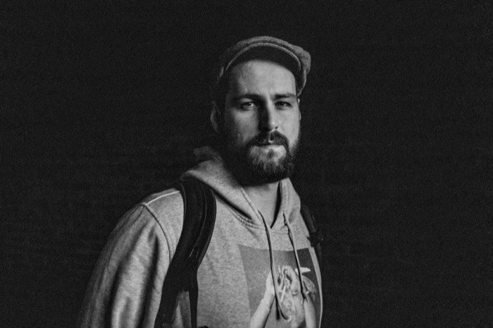 Bearded man in a cap, hoodie and backpack lit against a black background, gazing calmly at the viewer