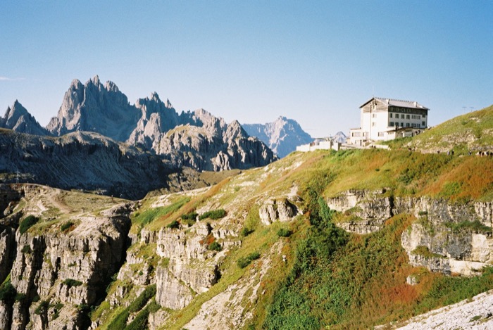 Large mountain refuge perched on a grassy ridge with serrated rock spires rising behind it