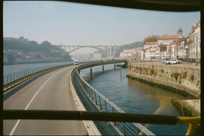 Riverside road curving toward an arched bridge, seen through the window frame of a moving vehicle