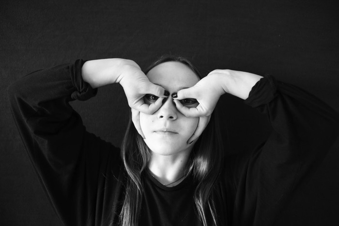 Girl against a black backdrop making binoculars with her curled hands over her eyes, long hair loose