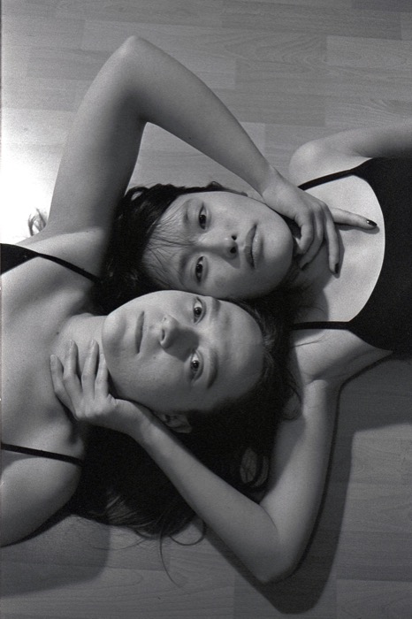 Two women lying head to head on a wooden floor, arms interlaced, both looking up at the camera