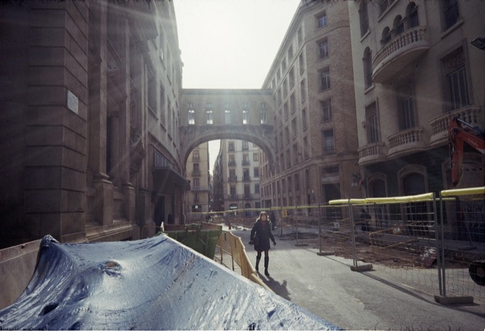Woman walking a barricaded street through roadworks toward a sunlit archway spanning stone buildings