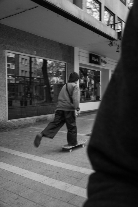 Skateboarder pushing off along an empty shopping street, past a shop sign reading "Black & White"