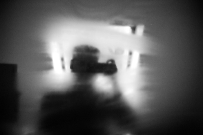 Blurred dark figure crouching in a bright empty room, motion smearing the scene into near-abstraction