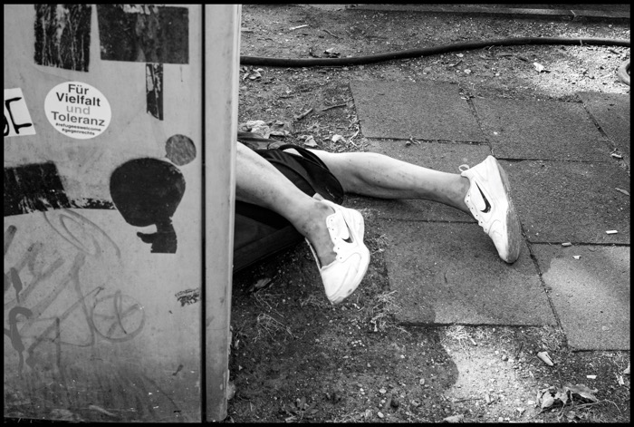 Bare legs in trainers stretched across a pavement behind a utility box stickered "Für Vielfalt und Toleranz"