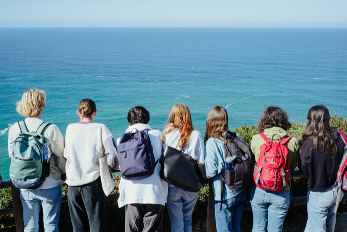 Seven people with backpacks lined up at a clifftop railing looking out over the open ocean