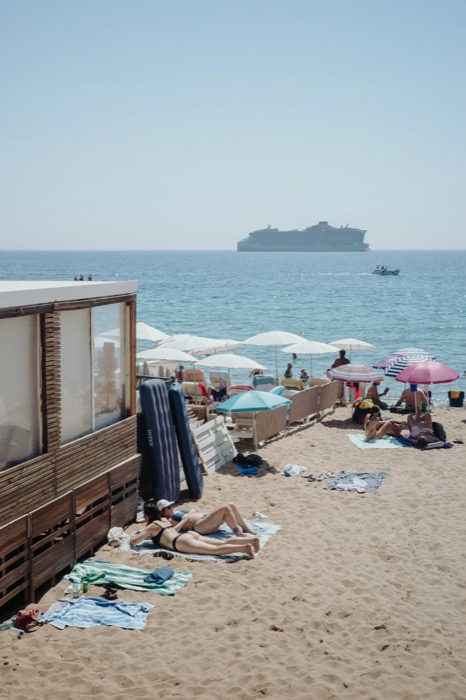 Sunbathers on towels among parasols on a city beach, a cruise ship anchored on the horizon