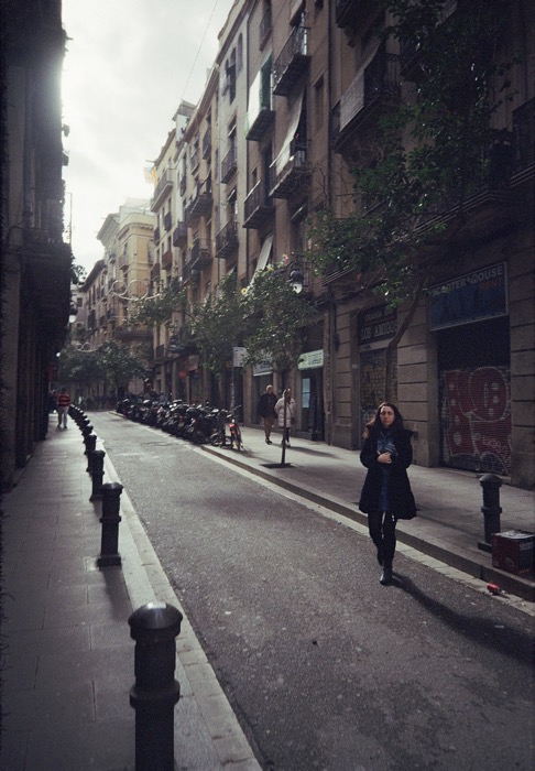 Woman in a dark coat walking up a narrow city street lined with balconies and parked scooters