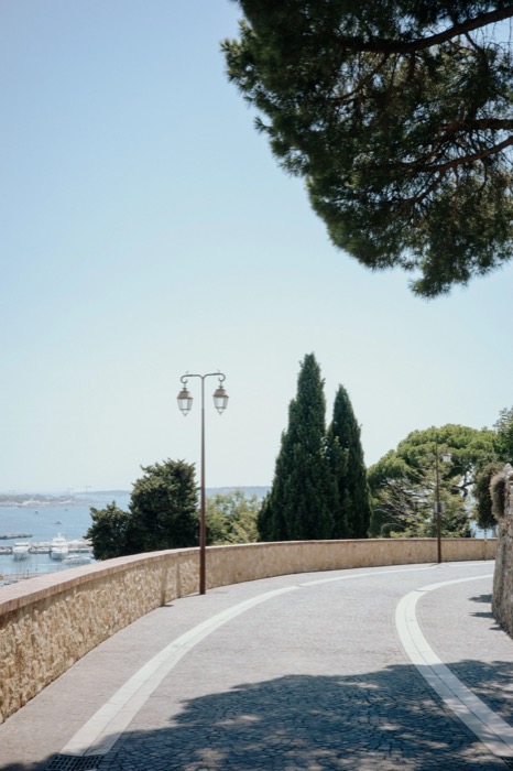 Empty curving cobbled road behind a stone wall, twin lamp post and cypresses above a marina