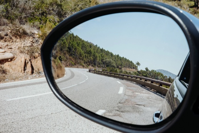 Car wing mirror reflecting the curve of a coastal road lined with pines and crash barrier
