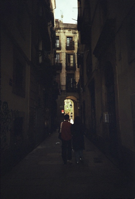 Two people walking away down a dark narrow alley toward a sunlit archway between old balconied houses